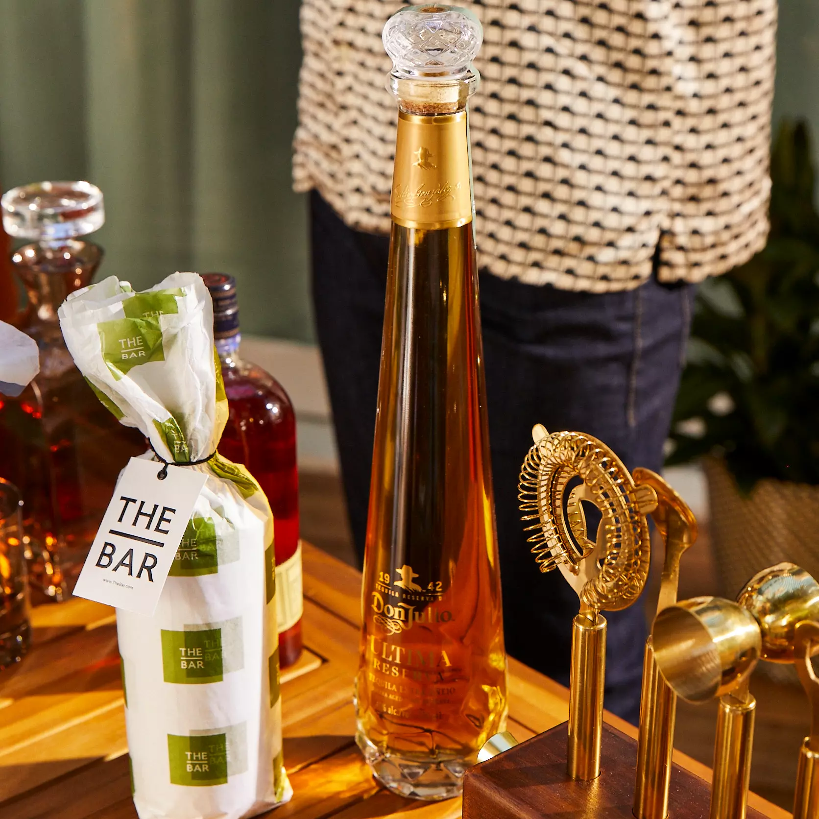 A tall, elegant bottle of Don Julio 1942 tequila stands on a wooden bar next to other liquor bottles and gold bar tools.