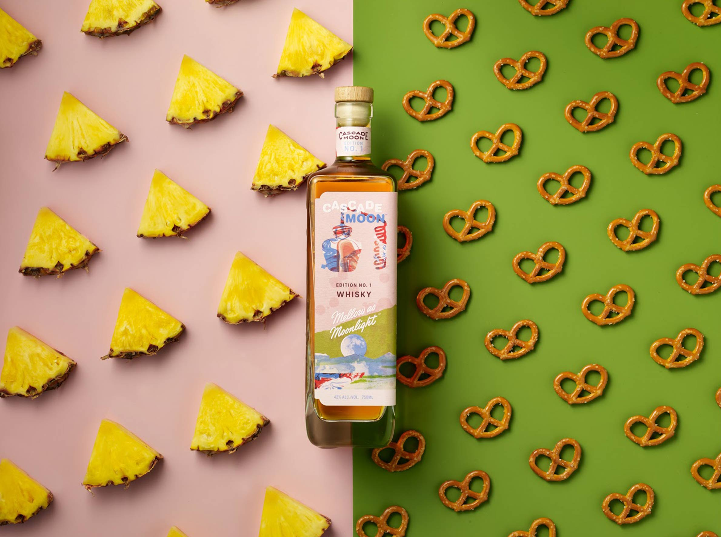 A bottle of Cascade Moon whisky is placed on a split background, with pineapple slices on the left and pretzels on the right.