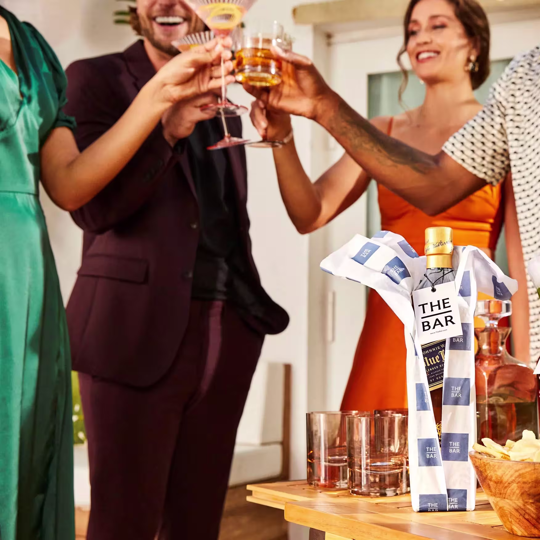 Four people clink glasses at a gathering, while a wrapped bottle labeled The Bar sits on a nearby table with empty glasses and a bowl of snacks.