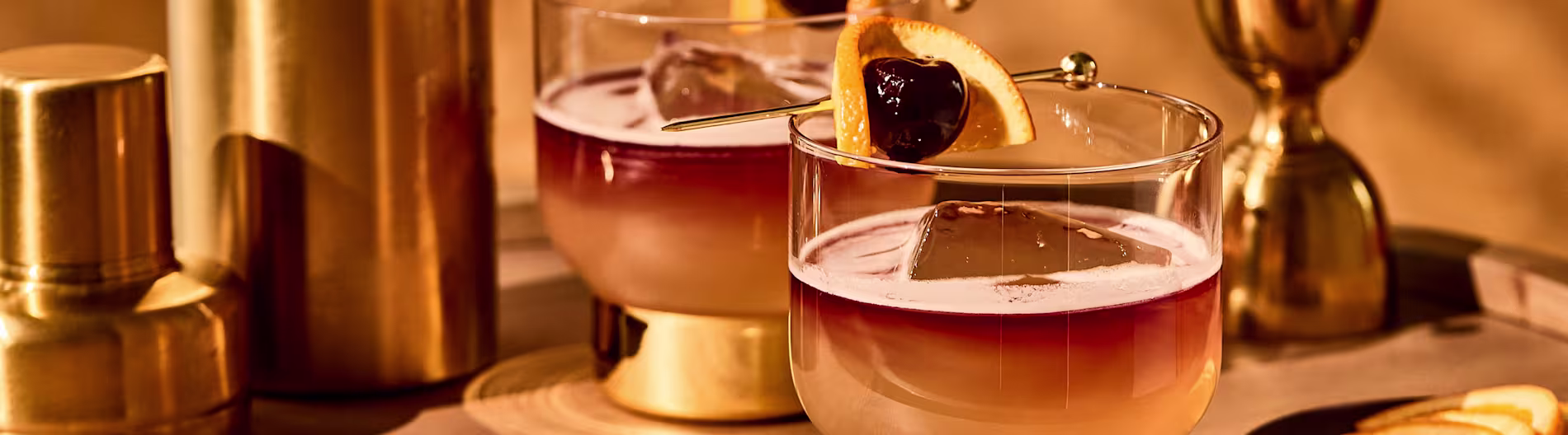 Barware în ton aurit alături de cocktailuri garnisite cu coajă de portocală și cireșe pe o suprafață elegantă