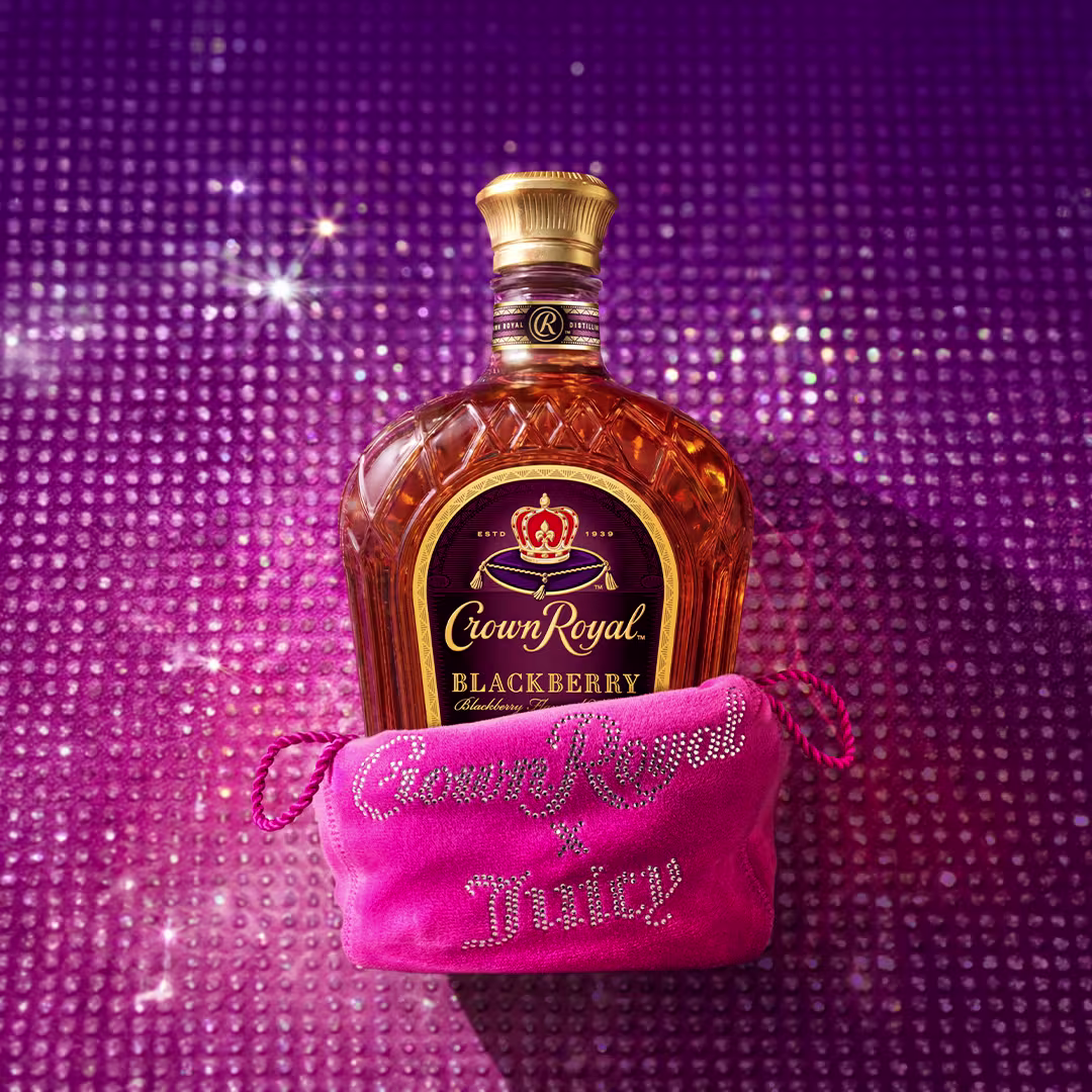 Crown Royal Blackberry whisky bottle displayed behind a bright pink Juicy Couture pouch embellished with rhinestones, set against a sparkling purple and pink backdrop.