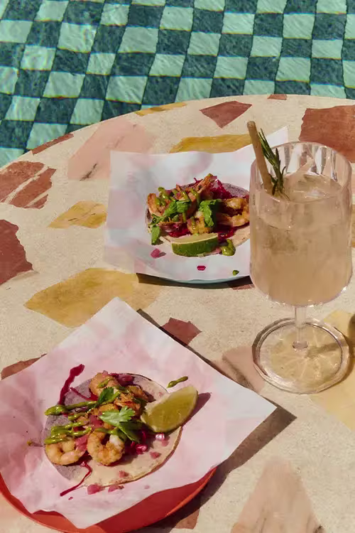 Mesa en Tope con tacos de camarón, lima, hierbas frescas y cóctel en copa alta, junto a una piscina con mosaicos azules.