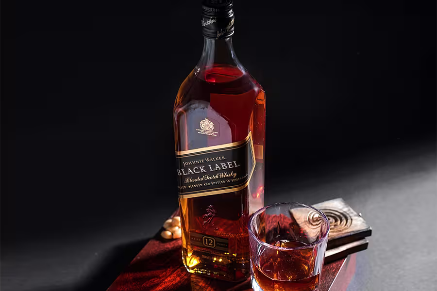 A bottle of Johnnie Walker Black Label whisky sits beside a glass of whisky on a wooden surface, with a dark background.