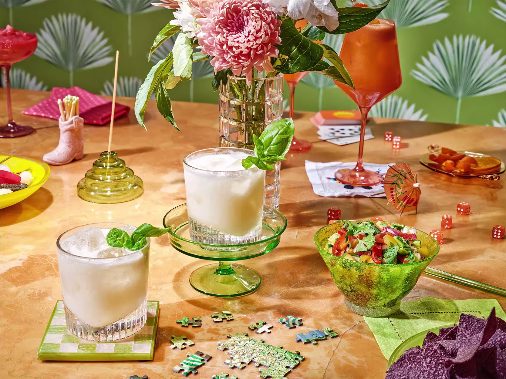 Two creamy cocktails with basil garnish on a table set with a salad, flowers, a cocktail, snacks, dice, playing cards, a puzzle, and bright green patterned wallpaper.