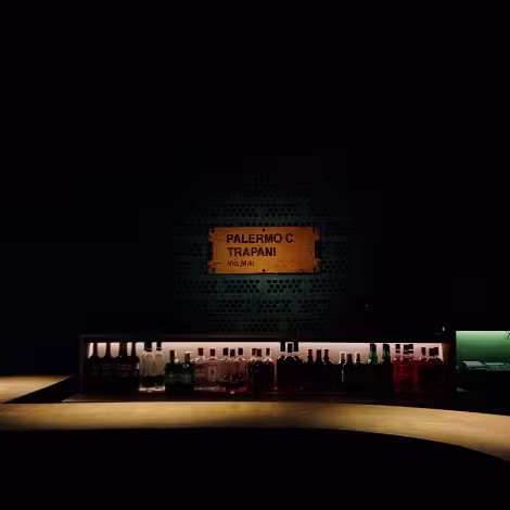 Una barra con botellas de varios licores en el fondo, ligeramente desenfocadas. Sobre las botellas, hay un letrero con las palabras “TRAPANI PALERMO” en letras mayúsculas.