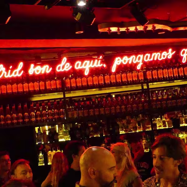 Bar animado con estanterías llenas de botellas y un neón rojo que dice "son de aquí, pongamos que". Ambiente vibrante y social.