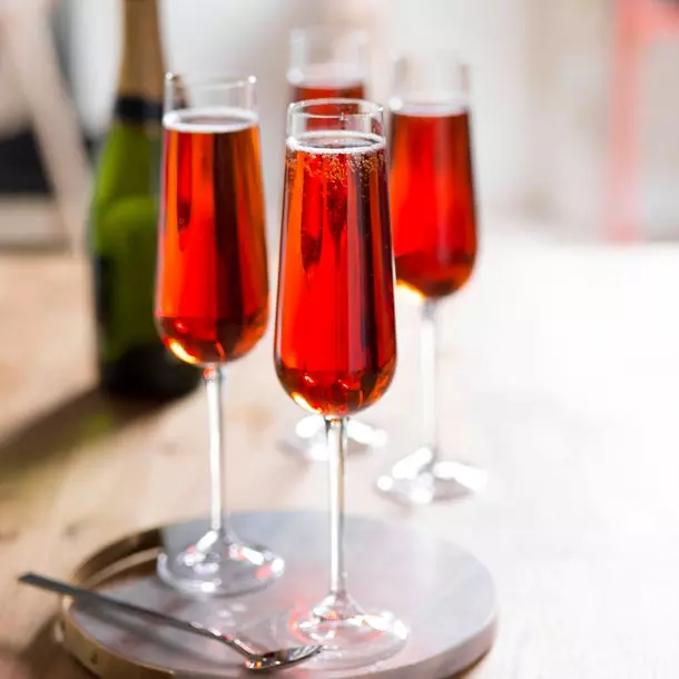 Four champagne flutes filled with a red cocktail are arranged on a round tray on a wooden table, with a bottle of sparkling wine in the background.