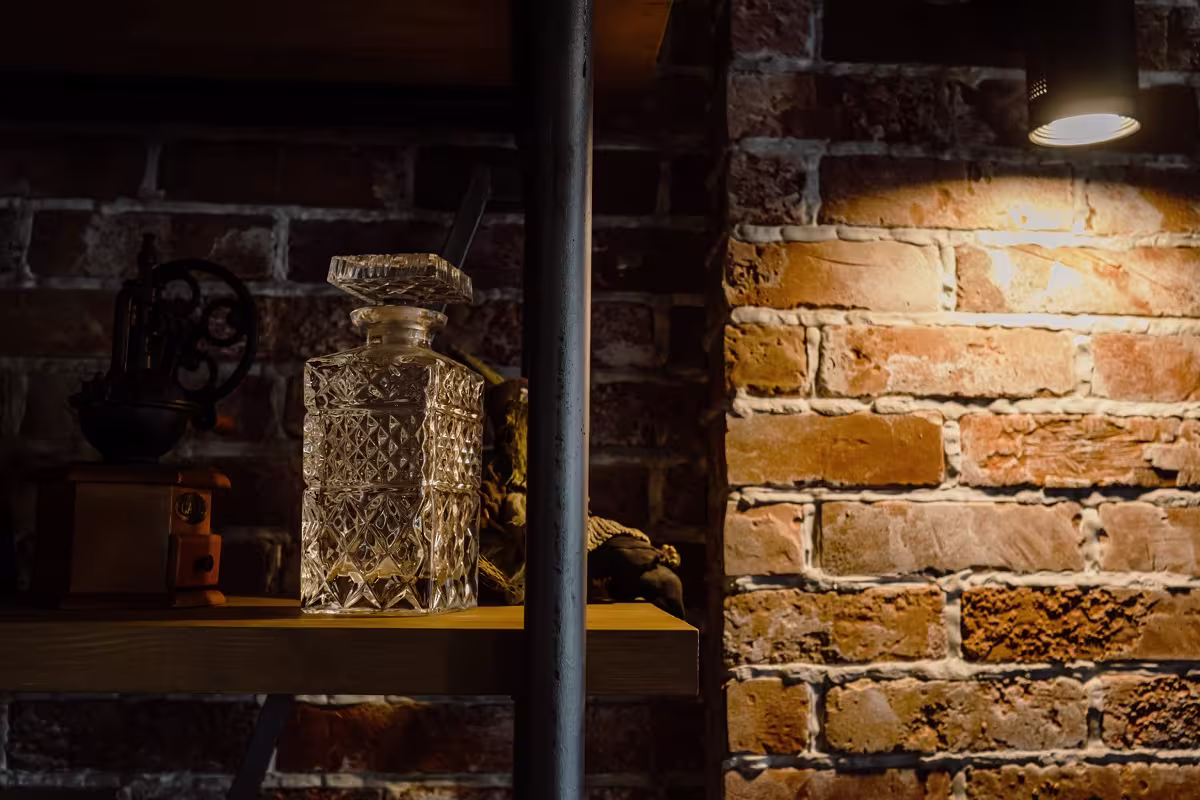 A glass decanter with a stopper sits on a wooden shelf against a brick wall, illuminated by a spotlight.