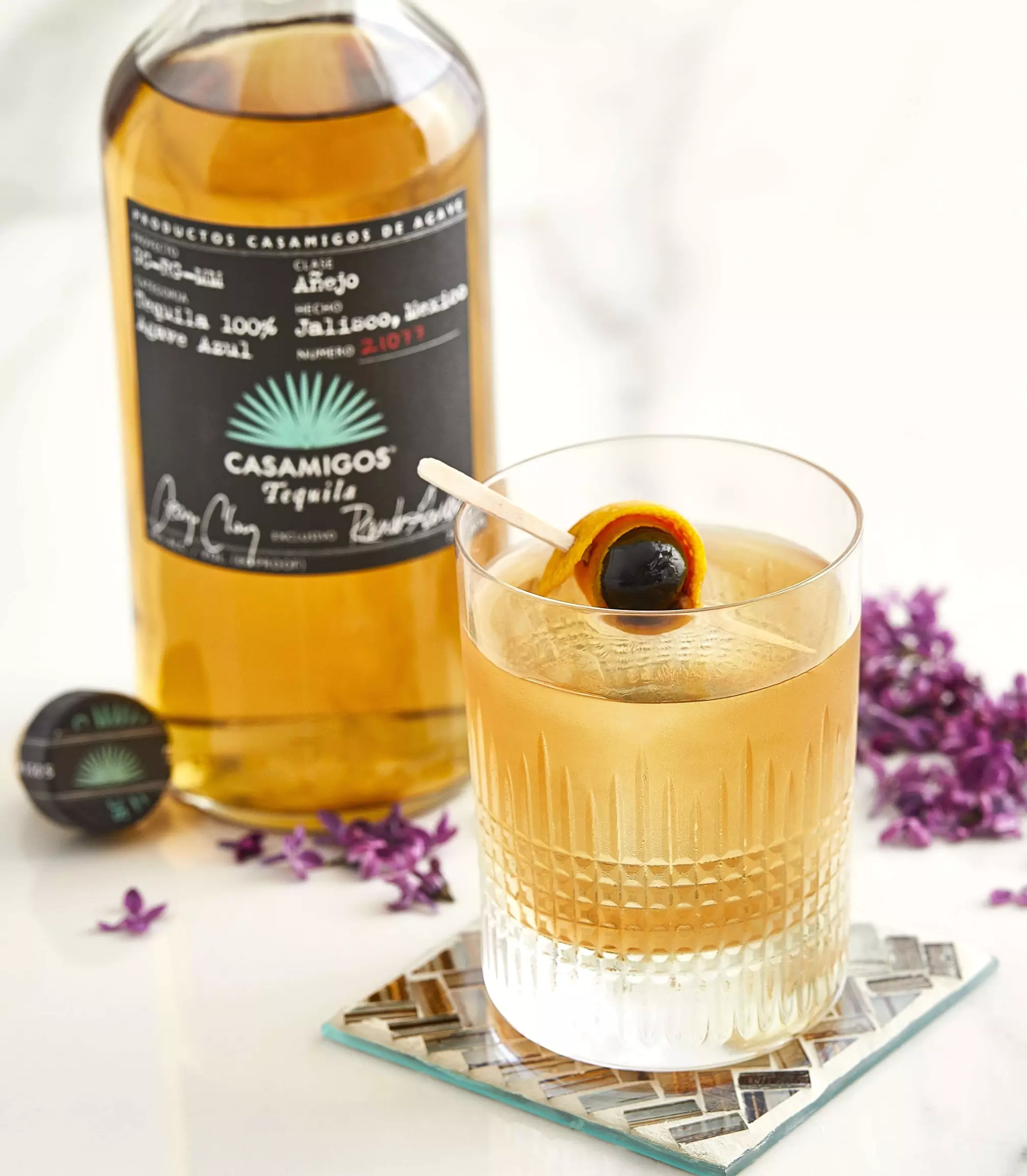 A glass of cocktail garnished with an orange peel and cherry sits in front of a bottle of Casamigos Añejo Tequila on a glass coaster, with purple flowers nearby.