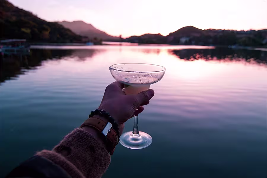 A person holds up a cocktail glass by a lake at sunset, with hills and water reflecting the sky in the background.