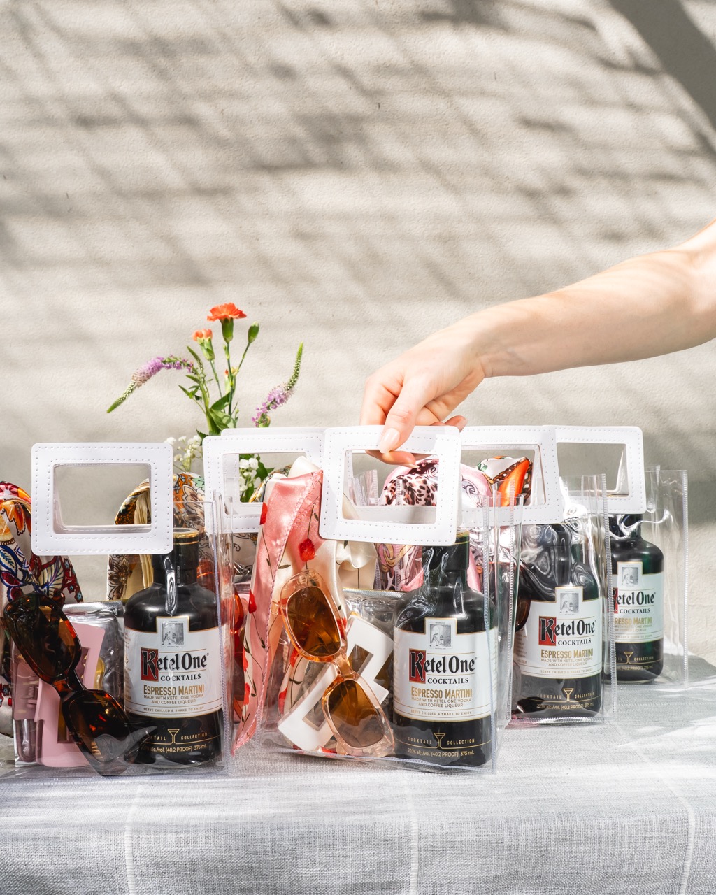 Clear bags containing Ketel One Espresso Martini bottles, sunglasses, and accessories are arranged on a table, with a hand picking up one bag; flowers are in the background.