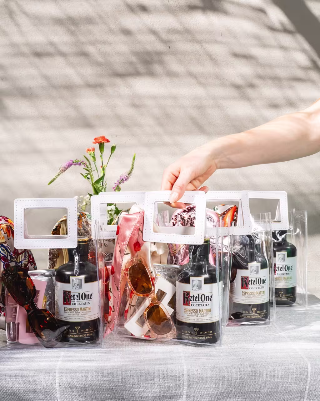 Clear bags containing Ketel One Espresso Martini bottles, sunglasses, and accessories are arranged on a table, with a hand picking up one bag; flowers are in the background.