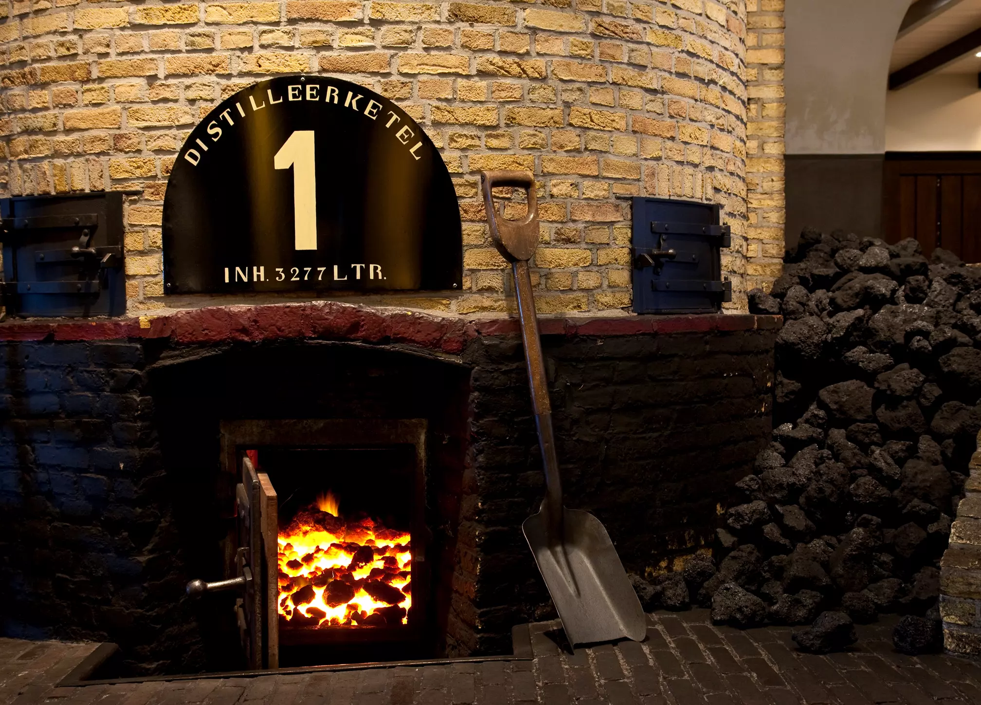 Large distillation kettle with an open fire at the base, a shovel leaning against it, and a pile of coal nearby. The sign reads DISTILLEERKETEL 1 INH. 3277 LTR.