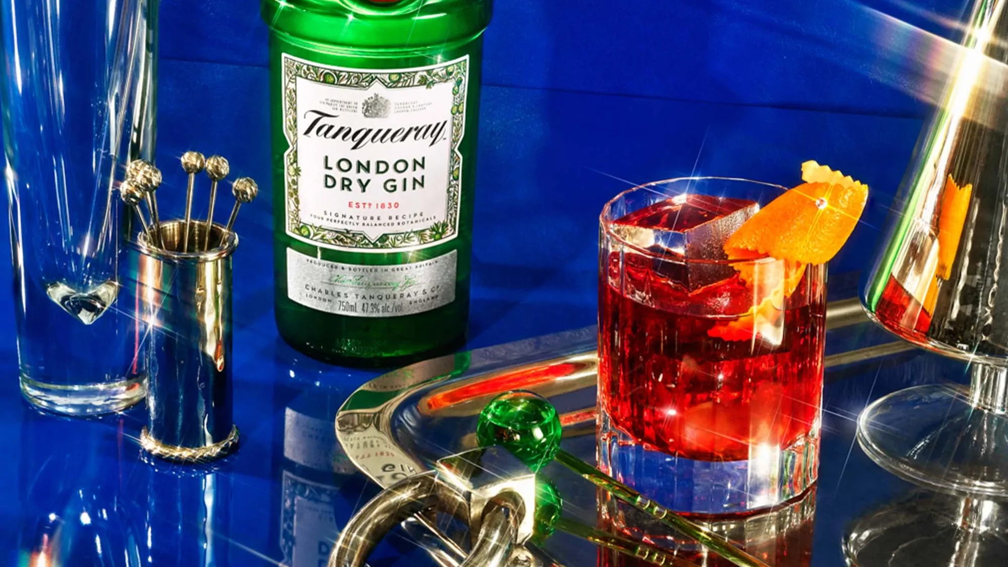 A bottle of Tanqueray London Dry Gin next to a glass of red cocktail with orange garnish, bar tools, and a tall empty glass on a reflective surface against a blue background.