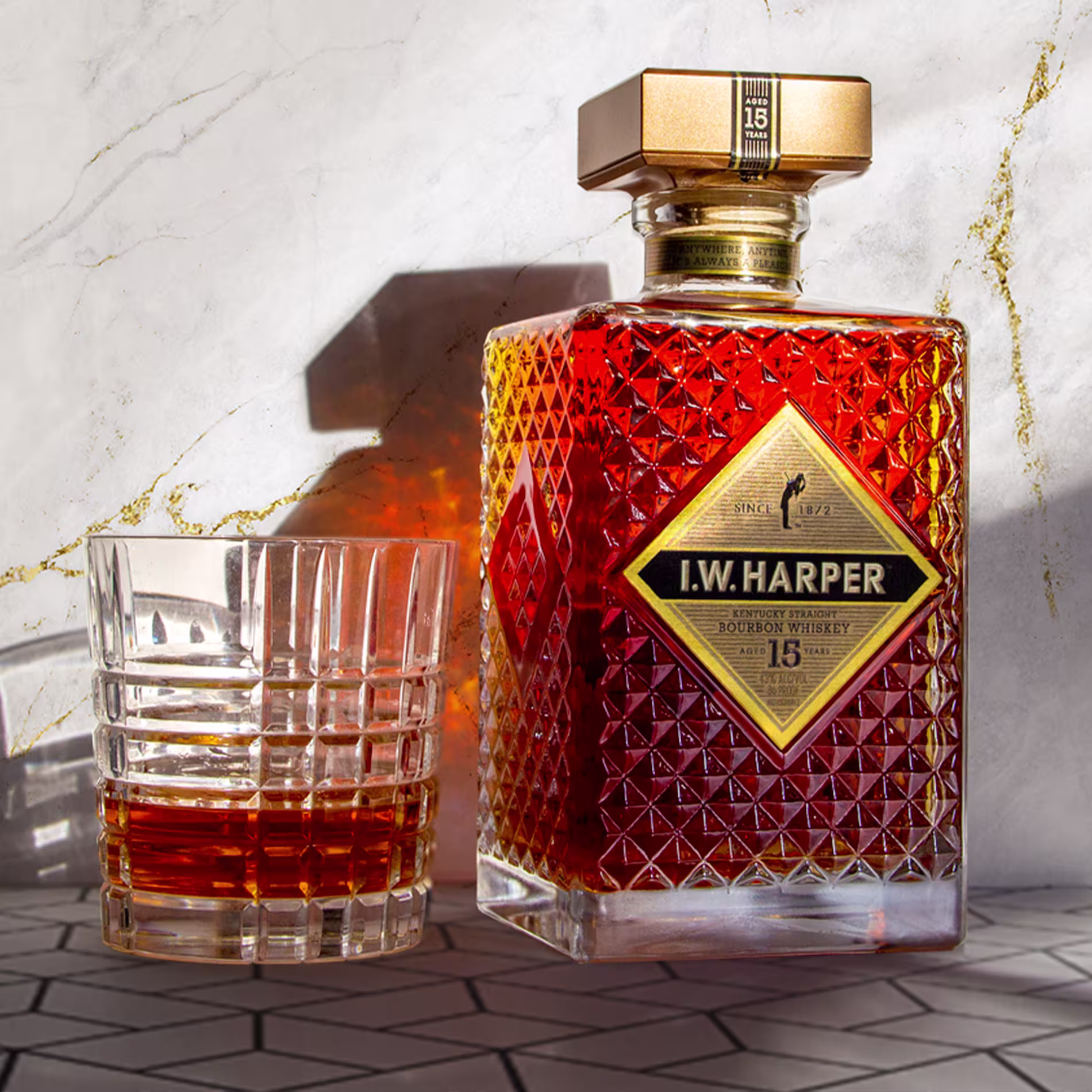 A bottle of I.W. Harper 15-Year-Old Bourbon Whiskey and a glass with whiskey sit on a patterned surface, both illuminated by sunlight.