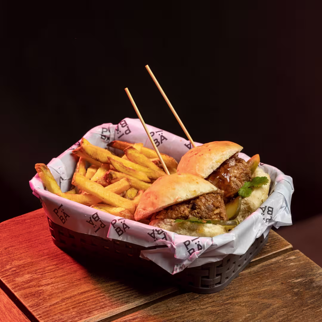 La imagen muestra una cesta que contiene una hamburguesa junto con una porción de papas fritas. Hay dos pinchos de madera que mantienen el sándwich unido. La cesta está forrada con papel que tiene el logotipo de PIBA
