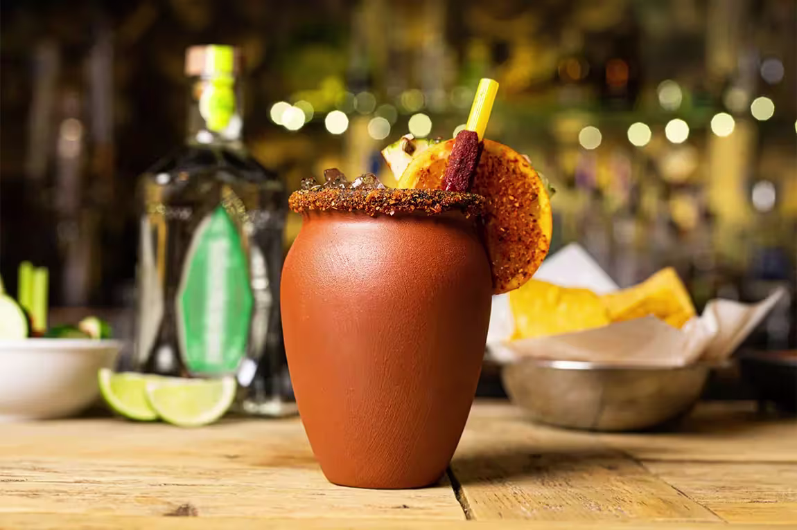 A clay cup with a chili-rimmed edge, filled with a dark drink and garnished with a spiced orange slice and tamarind stick, sits on a wooden table with tequila and lime in the background.