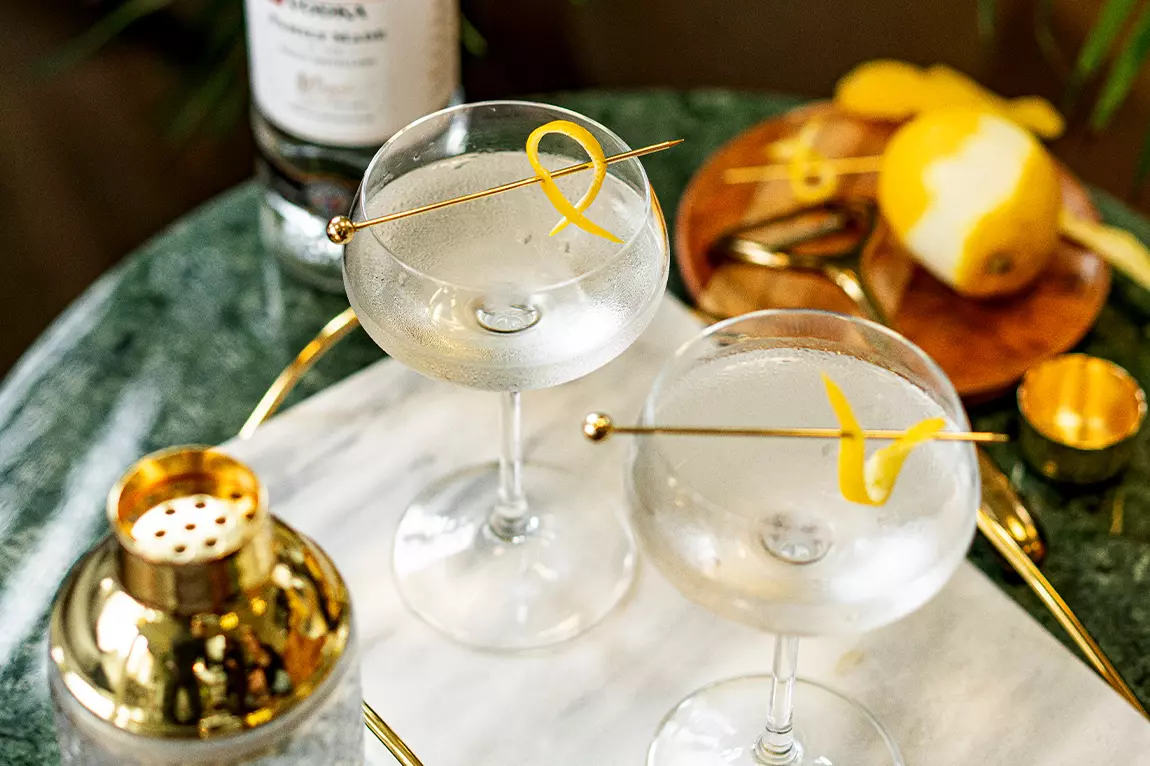 Two martini glasses with clear cocktails are garnished with lemon twists on a marble surface, next to a cocktail shaker, a bottle, and a peeled lemon.