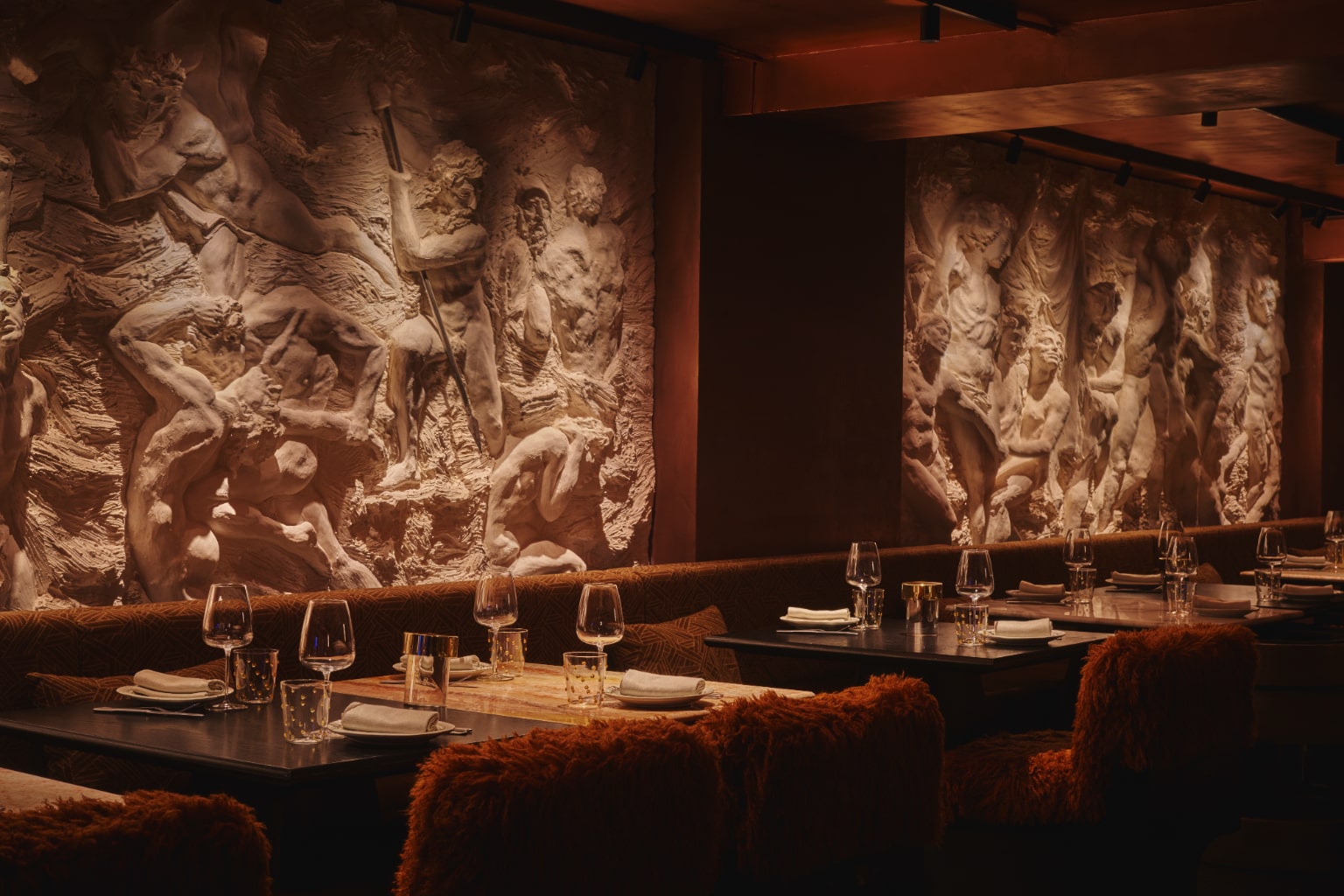 Interior del restaurante Rhudo con esculturas clásicas en relieve iluminadas cálidamente, mesas elegantes y sillas de terciopelo.