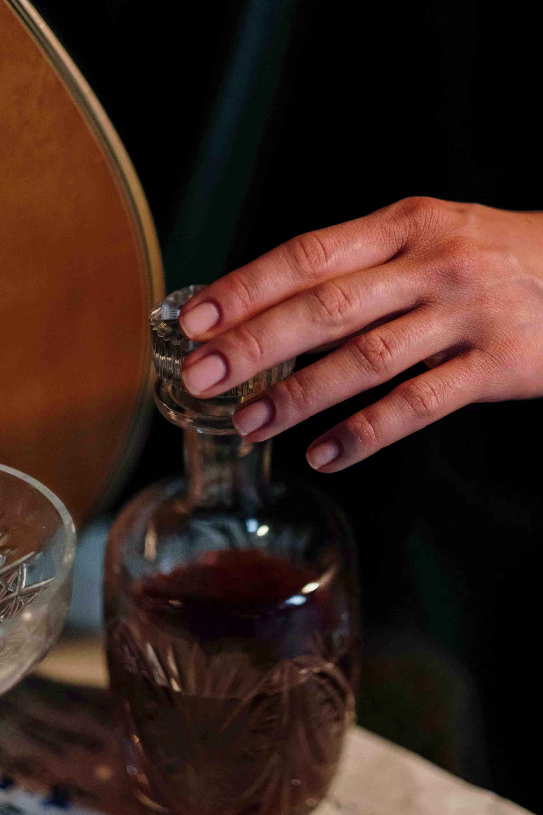 A hand is gripping the stopper of a glass decanter filled with a dark liquid, next to a crystal glass on a table.