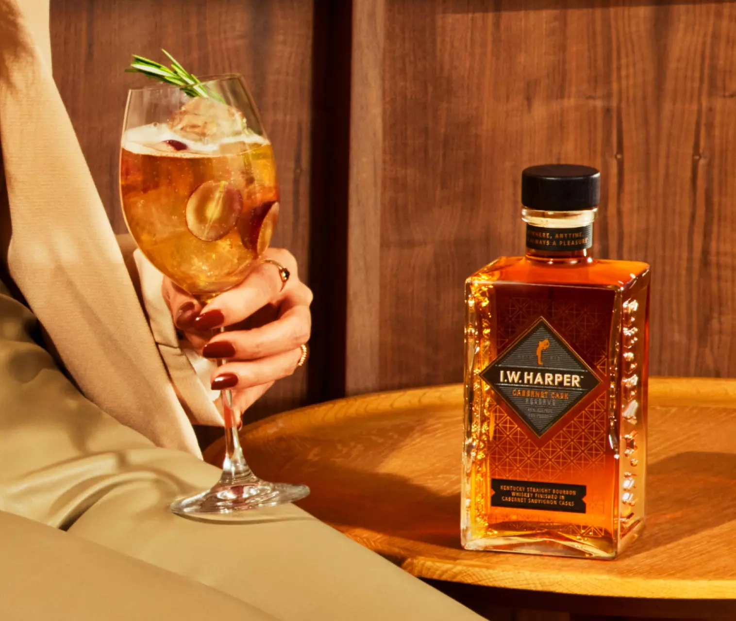 A person holds a cocktail glass with ice and a garnish next to a bottle of I.W. Harper Kentucky Straight Bourbon Whiskey on a wooden table.