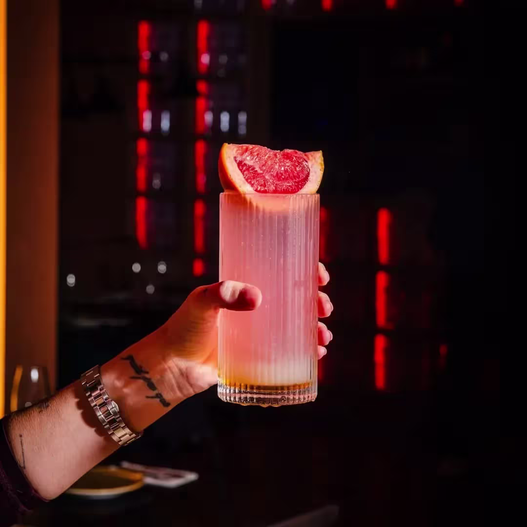 Una mano sostiene un cóctel rosa en vaso alto con textura, decorado con media rodaja de pomelo. Al fondo, luces rojas y ambiente nocturno.