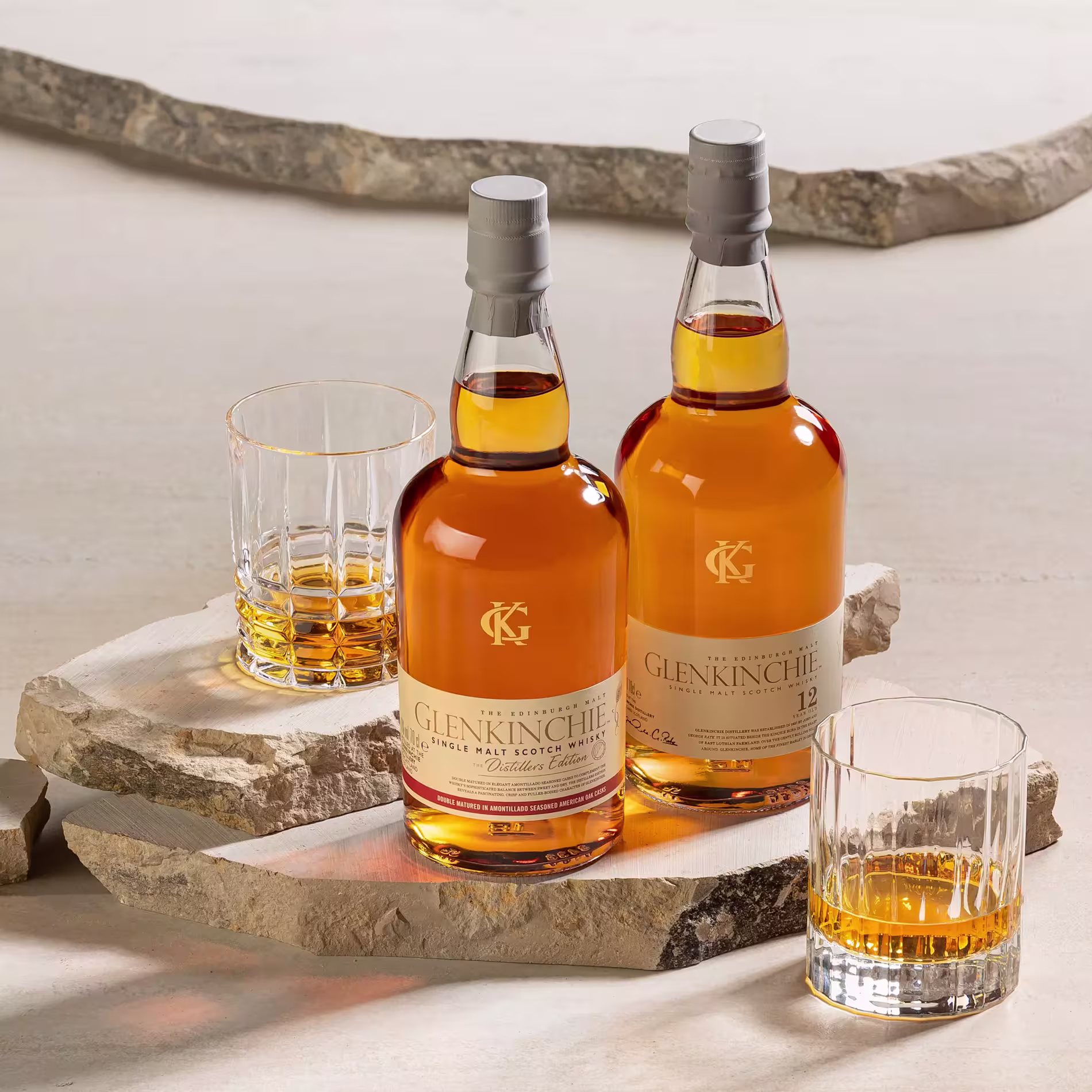 Two bottles of Glenkinchie whisky and two glasses with whisky are displayed on stone slabs against a neutral background.