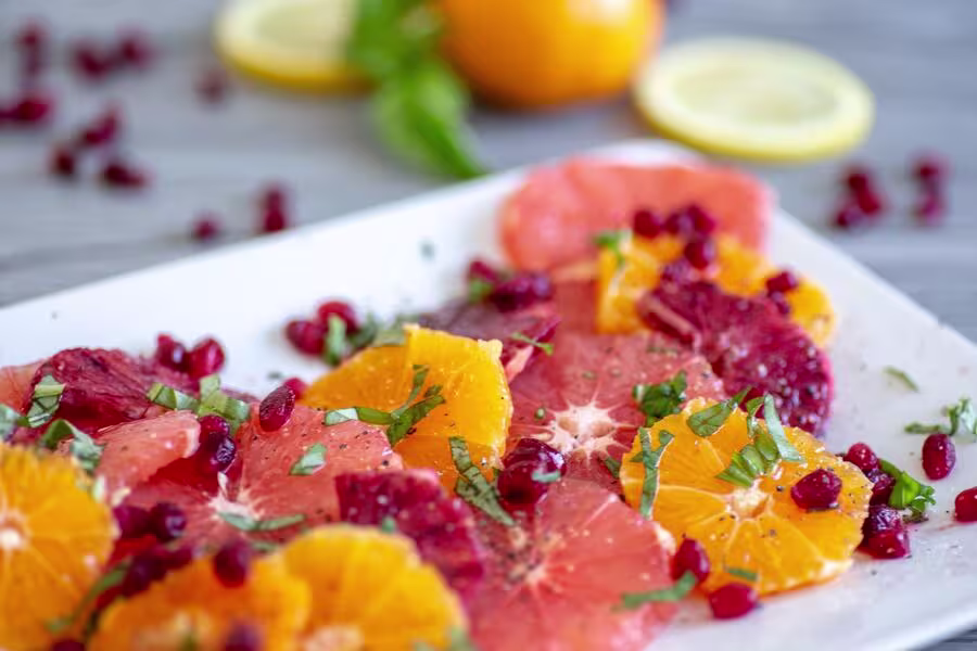 A plate of sliced citrus fruits, including oranges, blood oranges, and grapefruit, garnished with pomegranate seeds and chopped herbs.