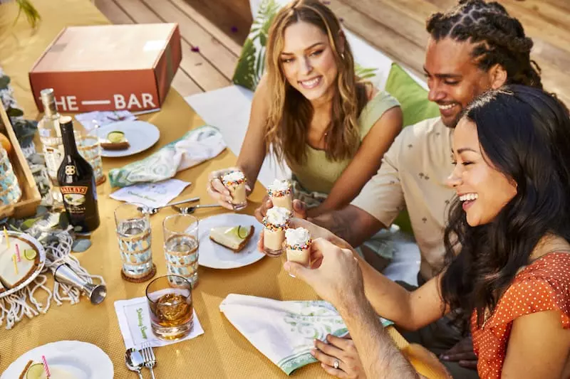 Three people sit at a table outdoors sharing drinks and desserts, smiling and enjoying a gathering with food, beverages, and decorations.