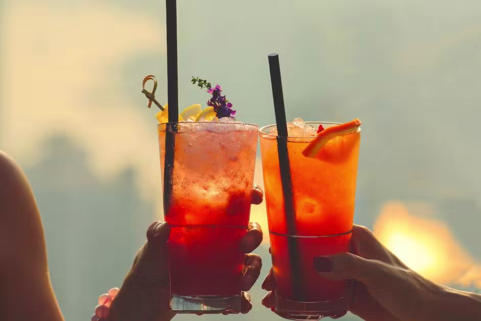 Two hands hold up glasses of red cocktails with ice, garnished with fruit and flowers, straws in each, against a blurred outdoor background.