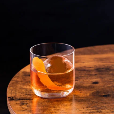 A glass of cocktail with ice and a lemon twist garnish sits on a white tiled bar counter with warm lighting in the background.