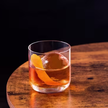 A glass of cocktail with ice and a lemon twist garnish sits on a white tiled bar counter with warm lighting in the background.