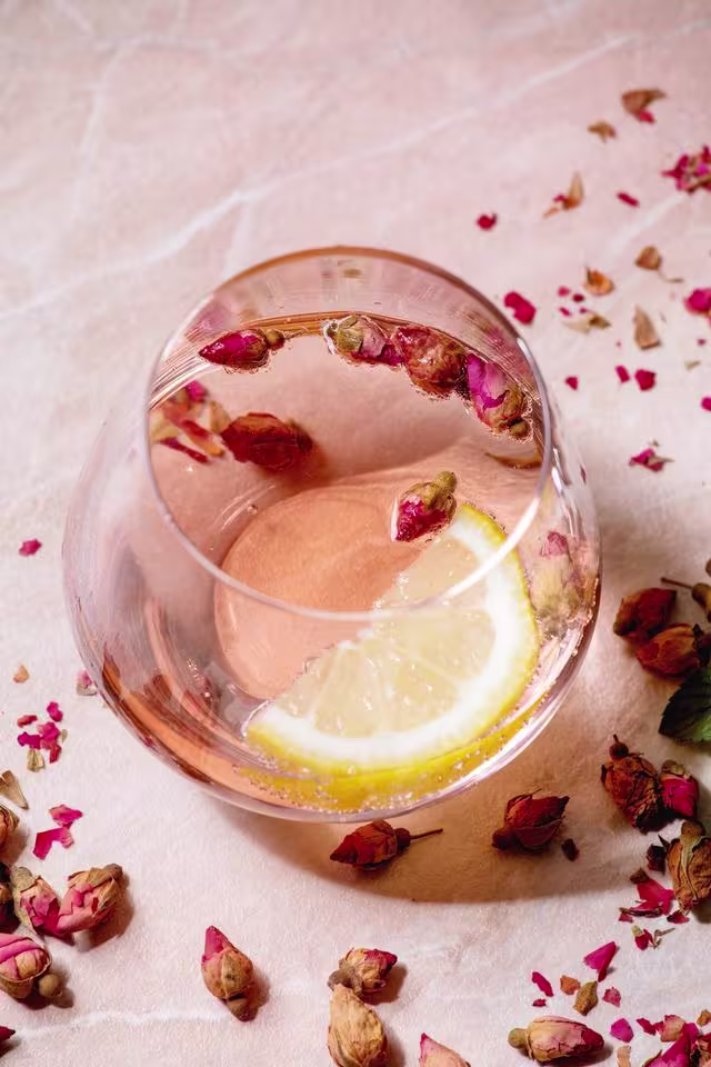 A glass of pink beverage garnished with dried rosebuds and a lemon slice, placed on a light surface with scattered dried rose petals.
