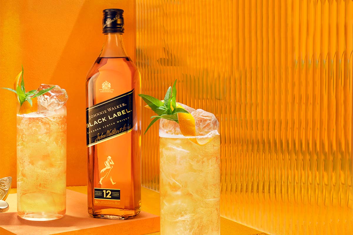 A bottle of Johnnie Walker Black Label whisky is placed beside two glasses filled with an orange cocktail, ice, and garnished with mint and orange peel, against an orange background.