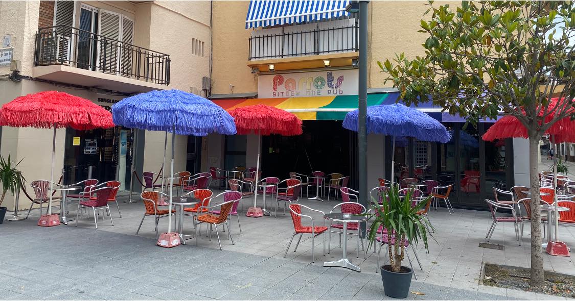 Terraza de Parrots, con sombrillas de colores vivos, sillas de metal en tonos vibrantes y un ambiente alegre en una plaza peatonal.