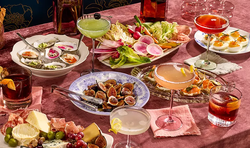 A table set with cocktails, oysters, figs, deviled eggs, assorted cheeses, charcuterie, and fresh vegetables on a pink tablecloth.