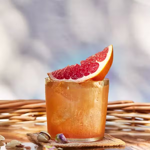 A glass of iced orange-colored drink garnished with a wedge of grapefruit sits on a coaster, with a wicker basket and pistachios nearby.