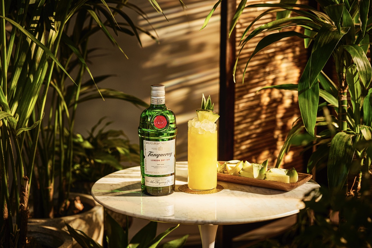 A bottle of Tanqueray gin, a yellow cocktail with ice and pineapple garnish, and a plate of pineapple slices on a round white table surrounded by green plants.
