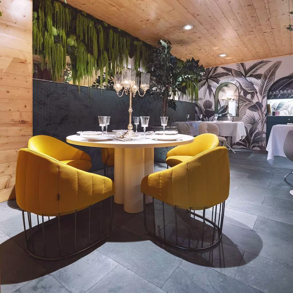 Espacio elegante con sillas amarillas, mesa redonda y candelabro. Pared con plantas colgantes y decoración tropical al fondo.