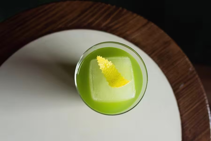 A green cocktail with a large ice cube and a lemon peel garnish, viewed from above, sits on a round table.