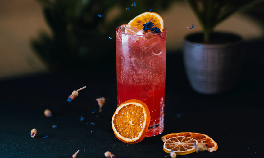A tall glass of red iced drink garnished with a dried orange slice and blue flower petals, with extra orange slices and petals scattered around on a dark surface.