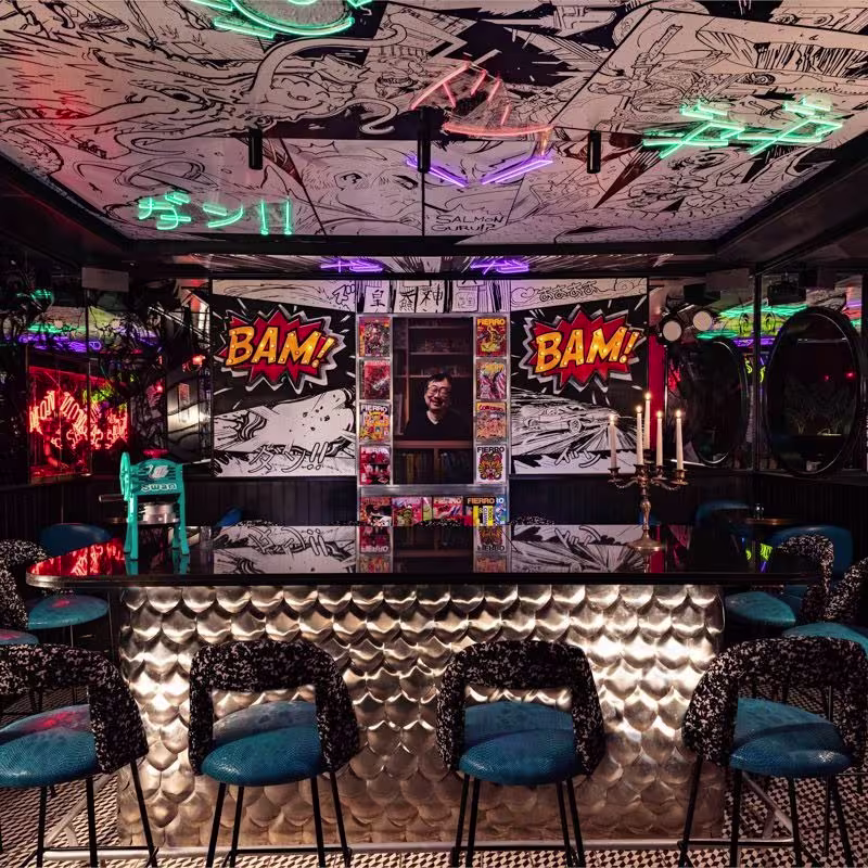 Barra metálica rodeada de sillas azules con respaldo estampado, decorada con murales de cómic y luces de neón en el techo.