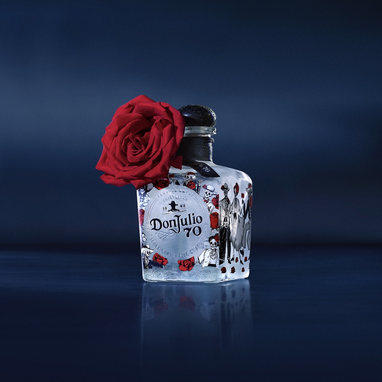 A bottle of Don Julio 70 tequila with a decorative label is adorned with a red rose attached to the neck, set against a dark blue background.