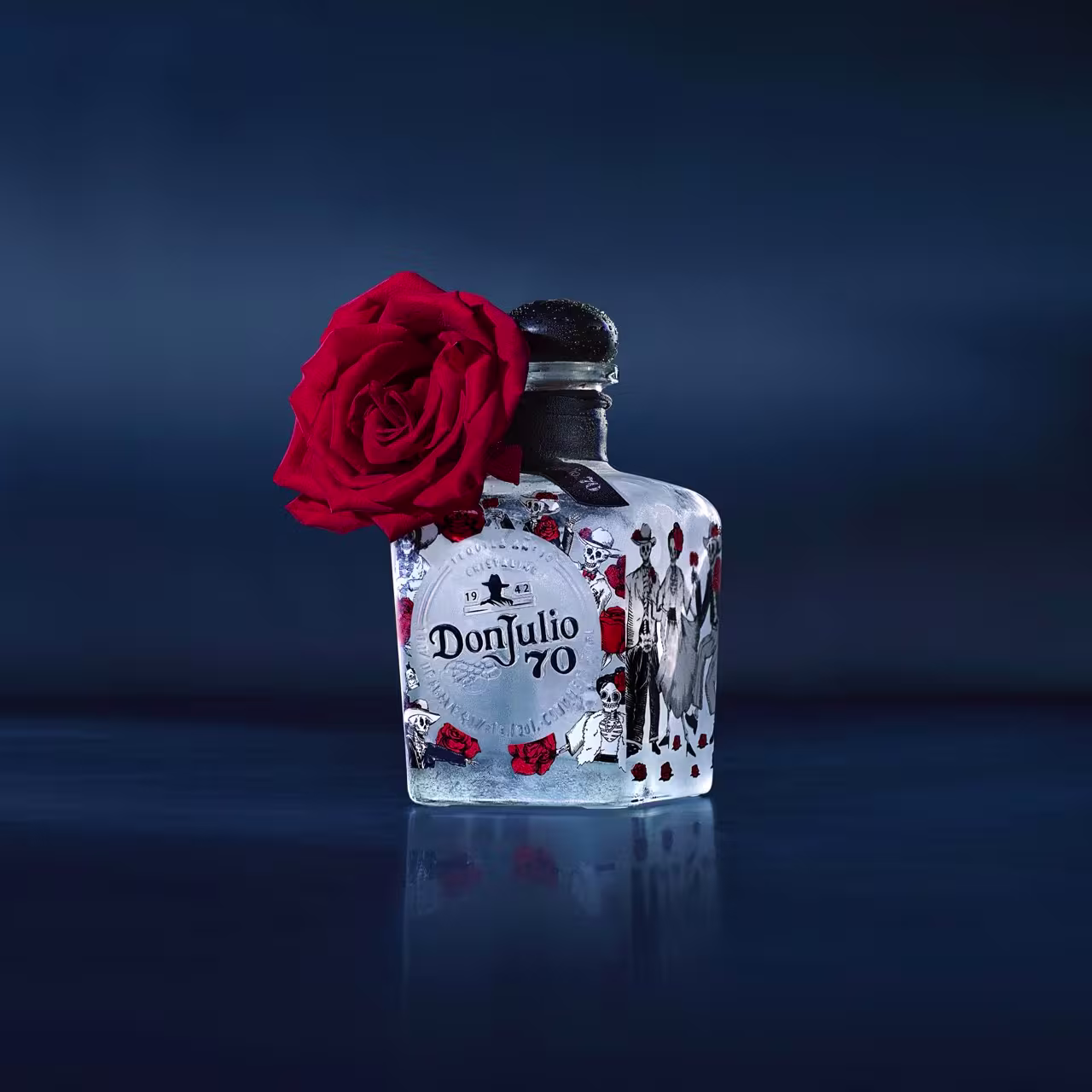 A bottle of Don Julio 70 tequila with a decorative label is adorned with a red rose attached to the neck, set against a dark blue background.