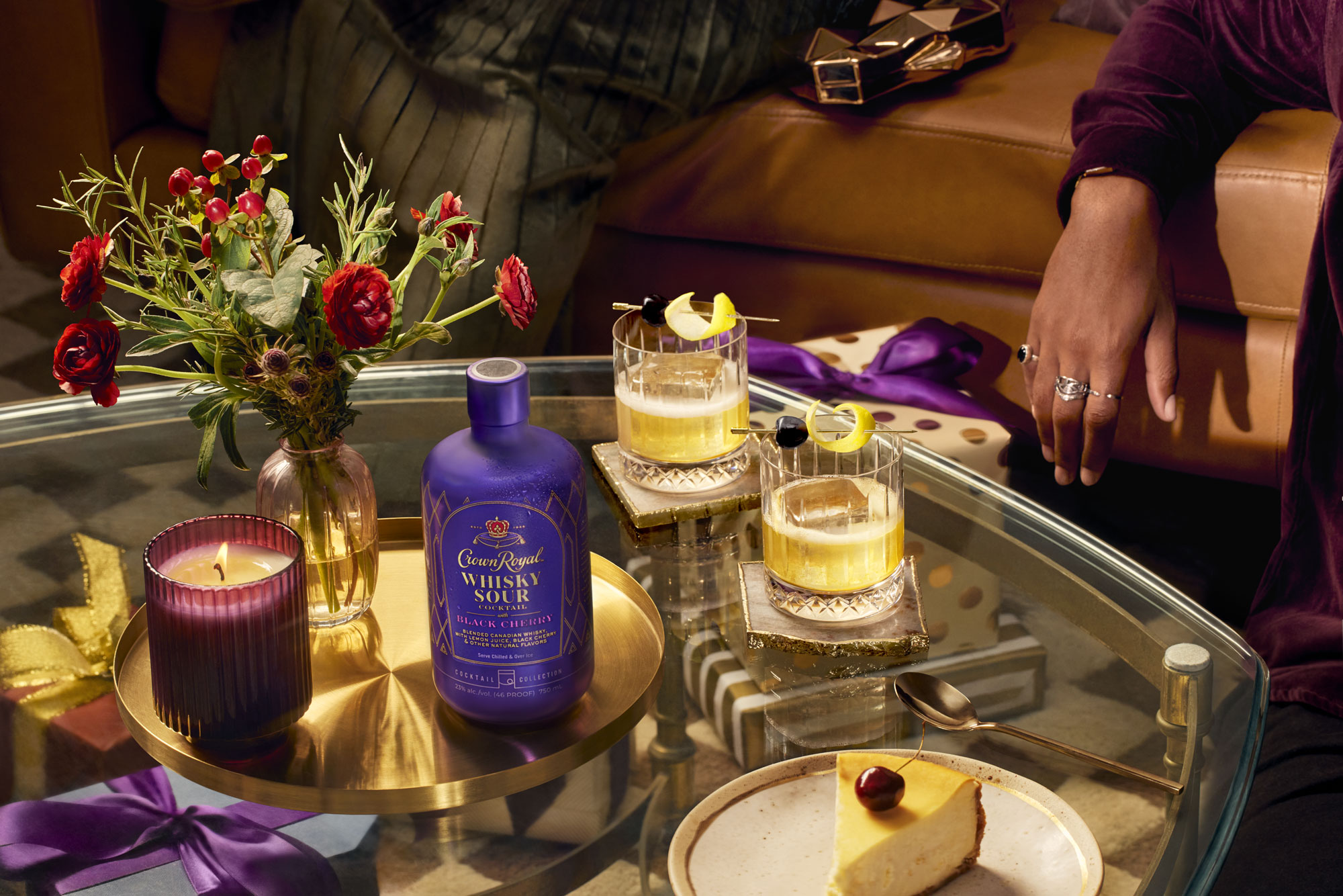A glass table with flowers, a lit candle, a bottle of Crown Royal Whisky Sour, two whisky cocktails, a slice of cheesecake, and a person’s arm resting on a chair.