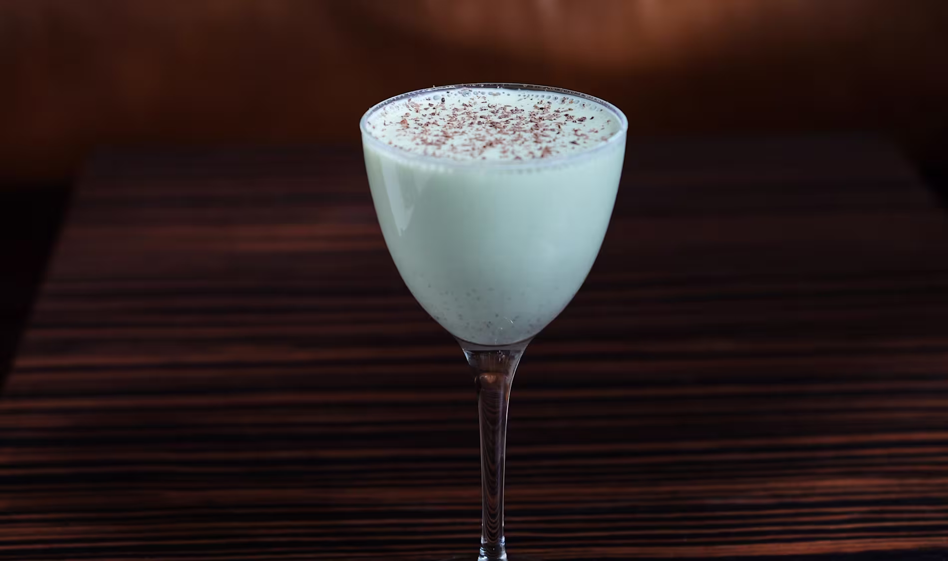 A stemmed glass filled with a creamy white drink, topped with a dusting of cocoa powder, sits on a dark wood surface.