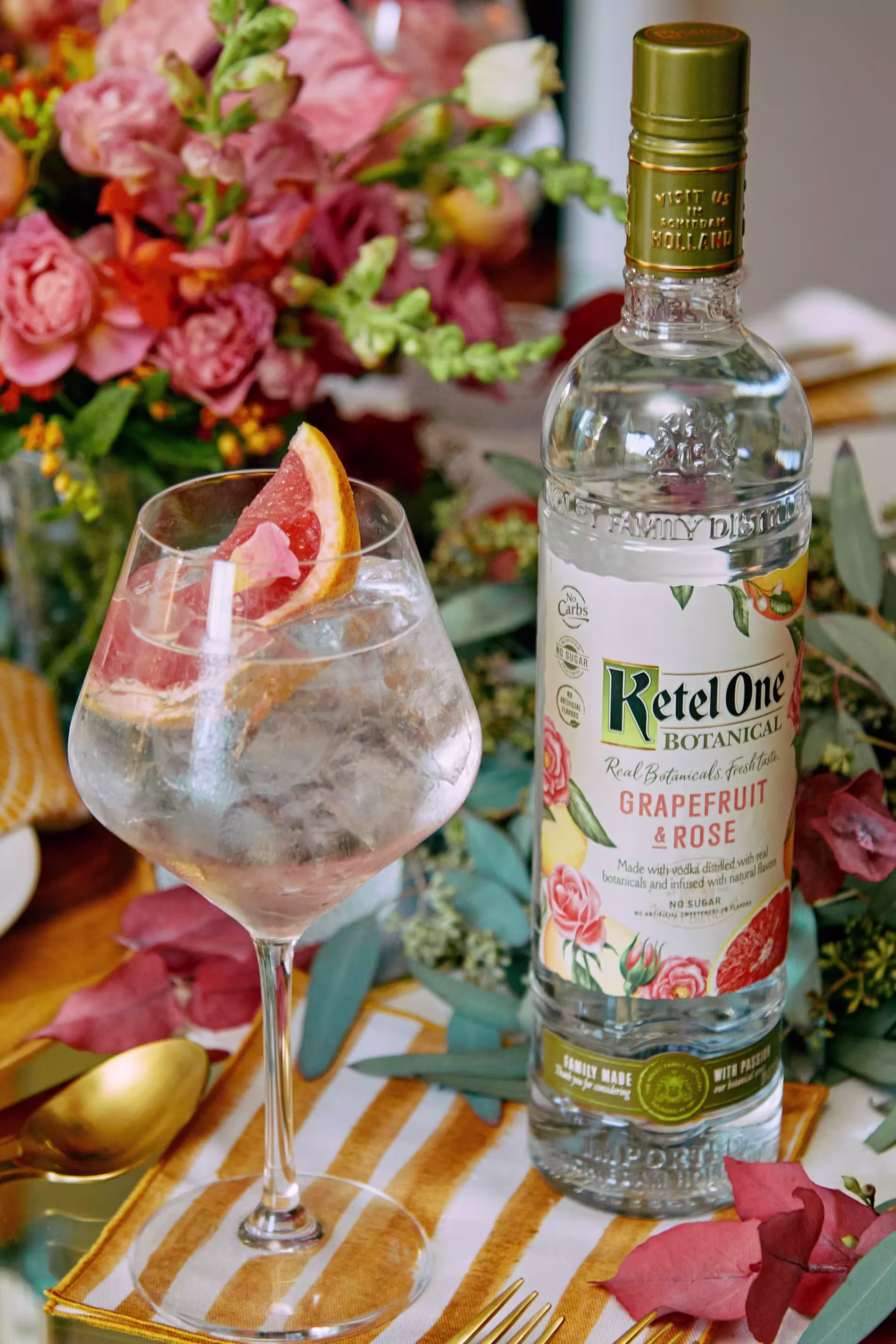 A bottle of Ketel One Botanical Grapefruit & Rose vodka stands next to a glass of cocktail with ice and a grapefruit slice, set on a decorated table with flowers.