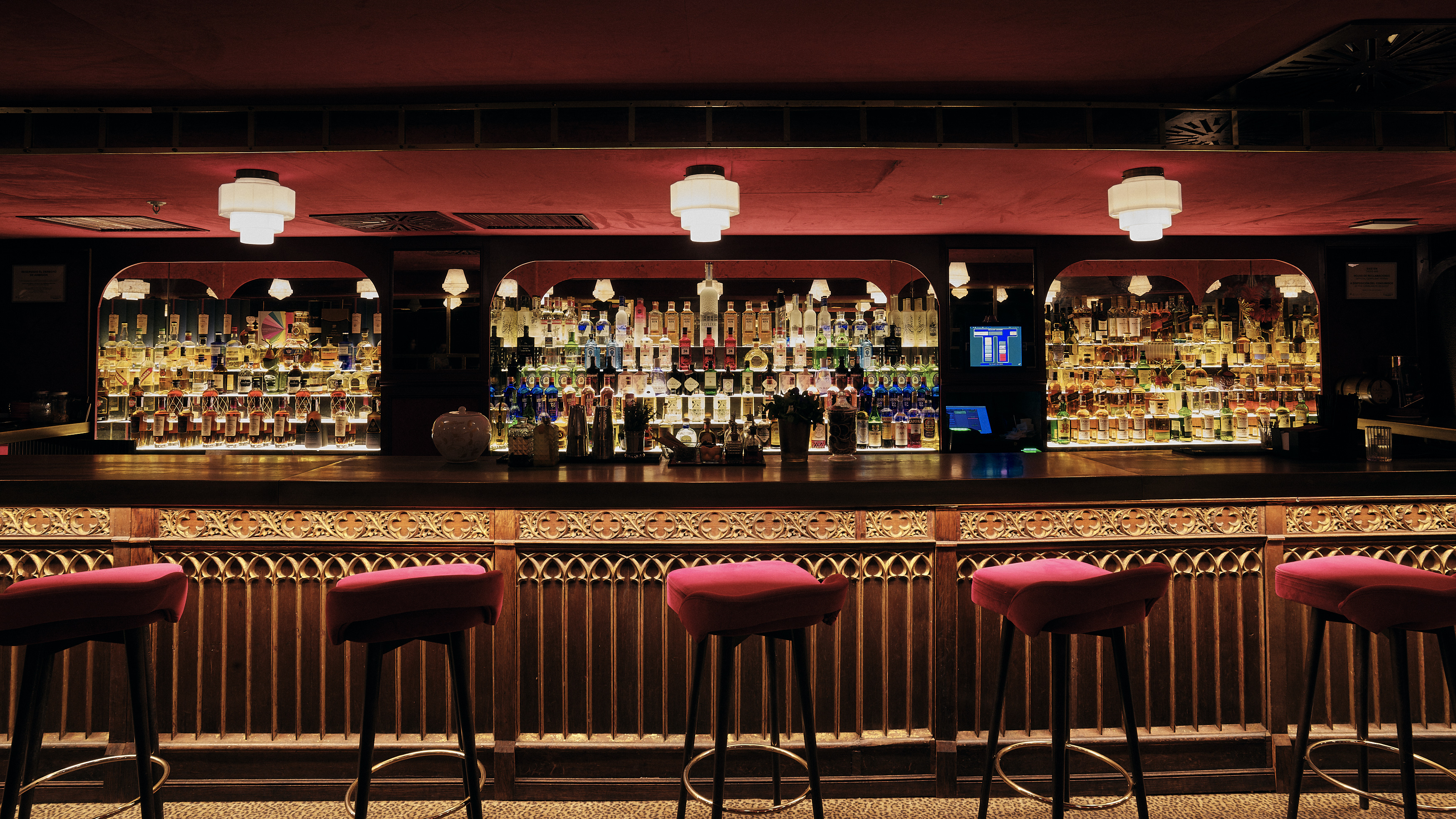 El interior de la coctelería Clandestino Cocktail Bar con una amplia selección de botellas en las estanterías de la pared del fondo, iluminadas con una luz cálida. Hay un largo mostrador con taburetes delante, la decoración es elegante y suave.