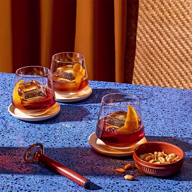 Three glasses of cocktails with orange peel sits on white coasters atop a blue tabletop, accompanied by a bowl of mixed nuts and a pen-shaped corkscrew.