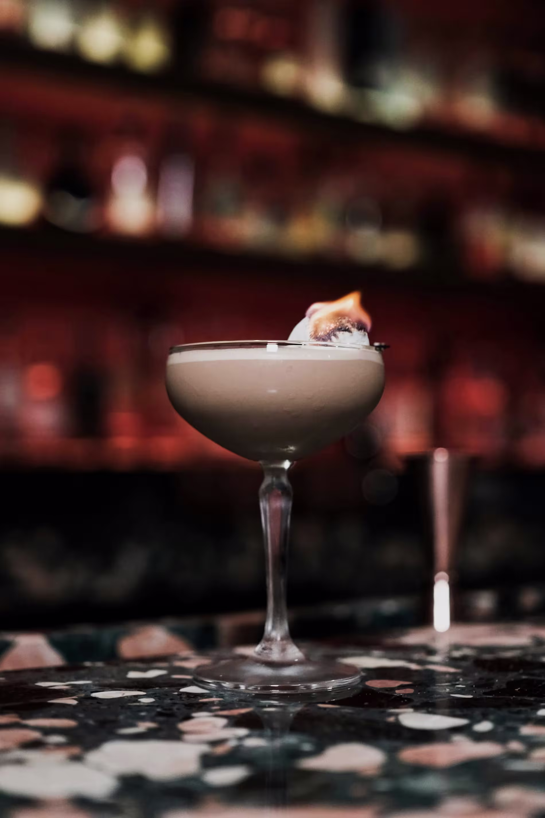 A creamy cocktail in a coupe glass sits on a marbled bar counter, garnished with a flaming marshmallow on a skewer.